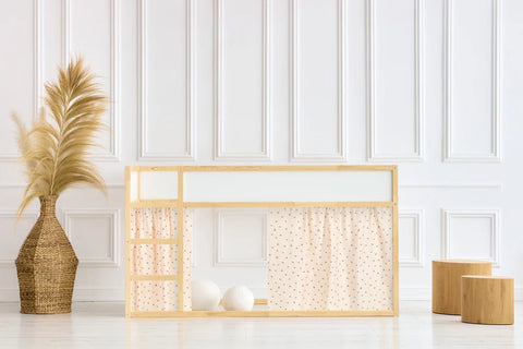 Minimalistisches Kinderspielhaus mit hellem Holzrahmen, gepunkteten Vorhängen und Stufen, neben einer großen Vase mit Pampasgras und zwei Holzhockern, vor einer weiß getäfelten Wand.