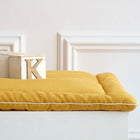 Eine KraftKids Wickelauflage Leinen gelb senfgelb mit weißer Paspel ruht auf einer weißen Fläche, auf der zwei Holzklötze (einer mit K) liegen. Zierleisten zieren die weiße Wand im Hintergrund.