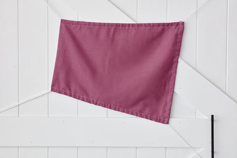 Eine einfarbig magenta-dunkle dreieckige Fahne von KraftKids, der Sonnensegel Leinen magenta dunkel, ist an eine weiße Holzwand mit diagonalen und vertikalen Paneelen gepinnt.