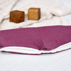 Der Kinderwagenmuff Leinen magenta dunkel von KraftKids, ein praktisches Kinderwagen Zubehör in lila und weiß mit schwarzen Druckknöpfen, ruht auf einer weißen Holzfläche neben einem cremefarbenen Tuch und zwei Holzklötzen.