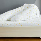 Ein KraftKids qualitäts Stillkissen Musselin im gelben Zitronenmuster liegt auf einem Holzbett mit weißer Bettwäsche und Decke, alles ergonomisch geformt und ordentlich zu einem V gefaltet vor einer grauen Wand und einem Hartholzboden.