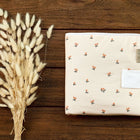 Eine gefaltete cremefarbene Babydecke mit kleinem Blumenmuster ruht neben getrockneten beigen Gräsern auf Holz, neben der KraftKids Reisewickelunterlage Musselin Kirschen - ideal für das praktische Wickeln unterwegs.