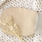 Ein Strampelsack Frühling Sommer Musselin beige von KraftKids mit einem hellgrauen Bündchen liegt auf einer dicken weißen Strickdecke, daneben ein kleiner Strauß getrocknetes beiges Gras.