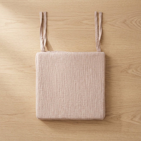 Ein hellrosa, strukturiertes KraftKids Sitzkissen für Stühle aus Musselin mit zwei Stoffbändern ruht auf einer hellen Holzoberfläche.