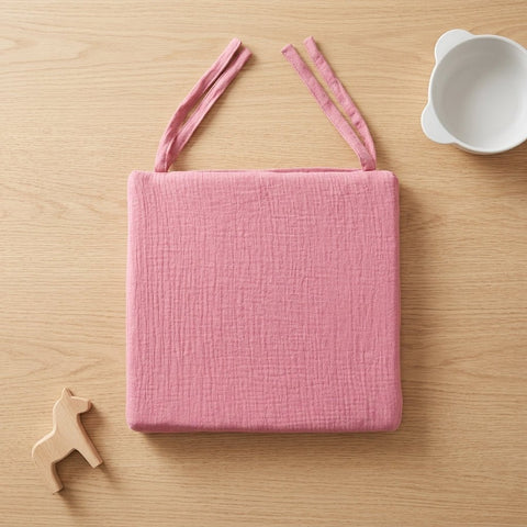 Ein pinkes KraftKids Sitzkissen für Stühle aus Musselin mit abnehmbarem Bezug und zwei Bändern liegt auf hellem Holz neben einer weißen Schale mit Bärenohren und einem kleinen Holzpferd.