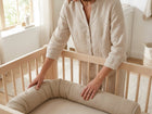 Eine Frau in beiger Kleidung stellt eine KraftKids Wickeltischrolle Musselin beige in ein helles Holzbett in einem sanft beleuchteten, neutral getönten Kinderzimmer.