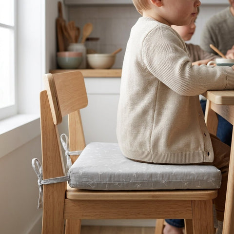 Ein kleines Kind sitzt an einem hölzernen Küchentisch auf einem Holzstuhl mit dem hellgrauen, gemusterten Sitzkissen für Stühle aus Musselin von KraftKids. Im Hintergrund erscheinen Regale mit Schüsseln und Küchenutensilien.