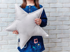Ein kleines Kind in einem blauen Kleid hält ein KraftKids Sternkissen Unigrau vor einer weißen Backsteinmauer, was es zu einer charmanten Ergänzung für jedes Kinderzimmer macht. Das Gesicht des Kindes ist nicht sichtbar.