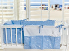 Ein weißes Kinderbett mit der KraftKids Betttasche abgerundete Dreiecke weiß auf Blau hängt in der Nähe, blaue Bettwäsche mit weißen geometrischen Mustern neben großen Fenstern in einem handgefertigten Kinderzimmer, Sonnenlicht strömt herein und gibt den Blick auf einen Sandstrand frei.