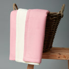 Eine KraftKids "Babydecke weiße Punkte auf Koralrosa" und eine schlichte weiße Fleecedecke sind über einem Weidenkorb auf einem Holzhocker drapiert und stehen vor einem neutralen grauen Hintergrund.