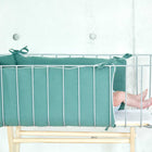 Die nackten Füße eines Babys schauen aus einem tealfarbenen, gepolsterten Bettchen mit Metallstäben hervor, das gemütlich in die weiche KraftKids Bettumrandung Nestchen Musselin vor einer hellgrauen Betonwand eingebettet ist.