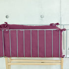 Ein KraftKids-Babybett aus Metall mit einem an den Ecken gebundenen Bettumrandung Nestchen Musselin purpur steht auf einem hellen Holzsockel und sorgt für wohlige Wärme vor einer weißen Betonwand.