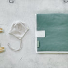 Ein Paar weiche gelbe Babyschuhe, ein weiß-grau gestreiftes Babymützchen, eine ordentlich gefaltete grüne Decke mit weißem Rand und die KraftKids Reisewickelunterlage Musselin nilblau auf einer hellgrauen strukturierten Oberfläche.