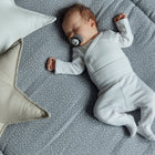Ein Baby in Weiß schläft friedlich auf der KraftKids Krabbeldecke Musselin grau Punkte, umgeben von Sternen und quadratischen Kissen, die mit weicher Polyestervlies-Füllung für Gemütlichkeit sorgen.