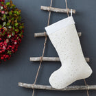 Die KraftKids Weihnachtssocke Musselin goldene Punkte auf Weiß hängt an einer rustikalen Holzleiter neben einem festlichen Kranz aus roten Beeren und grünen Blättern vor einem dunklen Hintergrund.