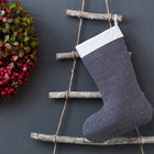 Eine festliche Weihnachtsdekoration mit roten Beeren und grünen Blättern hängt an einer grauen Wand neben der KraftKids Weihnachtssocke Musselin goldene Punkte auf Grau, die auf einer rustikalen Leiter aus Zweigen und Schnüren steht.