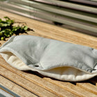 Der KraftKids Kinderwagenmuff Leinen leicht grünes Weiß, ein weicher und leichter Stoffbeutel in grünlichem Weiß, ruht auf Holzlatten mit einem belaubten Ast in der Nähe, während das Sonnenlicht sanfte Schatten auf die Szene wirft.