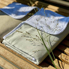 Zwei ordentlich gefaltete, helle KraftKids Wickelunterlage Leinen leicht grünes Weiß mit weißem Rand liegen auf einem Holzlattenrost, das Sonnenlicht wirft Schatten und zarte grüne Grashalme liegen über dieser gemütlichen Wickelunterlage.