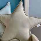 Ein Sternkissen Leinen leicht grünes Weiß von KraftKids, geformt wie ein Stern mit einem kleinen Stoffanhänger, ruht auf einem anderen hellen Kissen und einer blauen Wand und verleiht dem Kinderzimmer einen gemütlichen Stil.