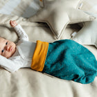 Ein Baby entspannt sich auf einem beigen Bett, trägt ein weißes langärmeliges Oberteil und den KraftKids Strampelsack Frühling Sommer Musselin goldene Punkte auf Petrol. Im Hintergrund sind sternförmige Kissen zu sehen.