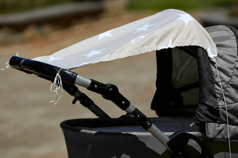 Nahaufnahme eines grauen Kinderwagens mit einem KraftKids Sonnensegel große weiße Sterne auf beigem Baumwollverdeck. Die Kanten haben ausgefranste Fäden, und der Hintergrund im Freien ist unscharf.