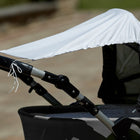 Eine Nahaufnahme zeigt einen Kinderwagen, über dessen Verdeck und Griffe das KraftKids Sonnensegel Karo grau drapiert ist, das den Kinderwagen effektiv vor der Sonne schützt. Der sanft verschwommene Hintergrund hebt den beschatteten Kinderwagen hervor.