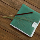Eine KraftKids Reisewickelunterlage Doppelkrepp Grün Jade mit weißem Rand und Etikett liegt auf einer Holzunterlage, die einem Notizbuch ähnelt. Ein brauner Stock und getrocknete Grashalme sind diagonal über die Wickelunterlage gelegt.