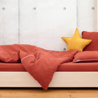 Ein ordentlich gemachtes Bett mit dem KraftKids Bettwäscheset Doppelkrepp Rot Herbstrot aus 100% Baumwolle, einem gelben Sternkissen und einem rostfarbenen Kissen auf einem leichten Holzrahmen - ideal für einen erholsamen Schlaf.