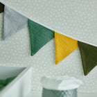Die Wimpelkette Doppelkrepp Grün Gelb Grau Herbstgrün von KraftKids mit farbigen Dreiecken hängt vor einer weiß-gepunkteten Wand und eignet sich perfekt als Kinderzimmer-Deko. Davor stehen ein weißes Tablett und ein strukturierter Behälter.