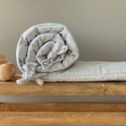 Eine KraftKids Bettumrandung Nestchen Cord Breitcord in Beige Taupe mit Schleifen liegt aufgerollt auf einer Holzbank neben drei glatten, runden Holzkugeln vor einer schlichten beigen Wand.