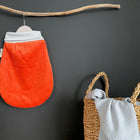 Ein KraftKids Strampelsack Frühling Sommer Cord Breitcord in Reinorange mit hellgrauem Deckel hängt an einem Holzast an einer dunklen Wand. Daneben hält ein Korb eine hellgraue Decke, die über den Rand drapiert ist.