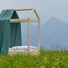 Ein einfaches Holzbett mit weißem Bettzeug und dem Bettbaldachin Musselin nilblau von KraftKids steht auf einer grünen Wiese mit Wildblumen, Bergen und einem wolkenverhangenen Himmel im Hintergrund.