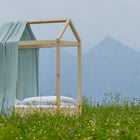 Das KraftKids Bettbaldachin Musselin mint Pusteblumen steht auf einem Holzrahmen mit mintfarbenen Akzenten, eingebettet in eine Wildblumenwiese mit Bergen - eine ideale Kinderzimmer Dekoration.