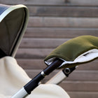 Eine Nahaufnahme des KraftKids Kinderwagenmuff Doppelkrepp Grün Herbstgrün, der an einem Kinderwagenlenker befestigt ist und die Hände warm hält, vor einer Holzbohlenwand.