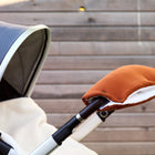 Nahaufnahme eines Kinderwagens mit grauem Verdeck und dem am Lenker befestigten KraftKids Kinderwagenmuff Doppelkrepp Rot Herbstrot vor dem Hintergrund eines Holzzauns.