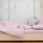 Ein helles Holzbett mit KraftKids Bettwäscheset Doppelkrepp Rosa aus 100% Baumwolle, inklusive einer handgefertigten Decke und Kissen - ein Stern in Hellgrau - steht an einer Betonwand in einem ungemachten Raum mit hellem Holzboden.