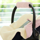 Nahaufnahme einer leeren schwarzen Babyschale mit der KraftKids Decke für Babyschale Winter Doppelkrepp Rosa aus weichem cremefarbenem und rosafarbenem Stoff mit Gurtschlitzen, die in einem Innenraum vor einem Fenster mit Grünpflanzen steht.