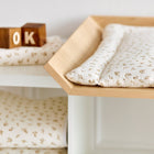 Die KraftKids Wickelauflage Musselin Zweige braun, mit zartem Zweigdruck und passender Decke, liegt auf dem Wickeltisch neben Holzklötzen, die auf einem weißen Regal OK buchstabieren.